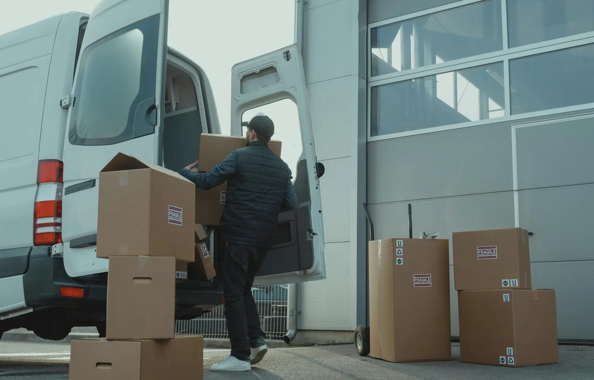 A delivery man unloading cardboard boxes from a van at a warehouse during the day.